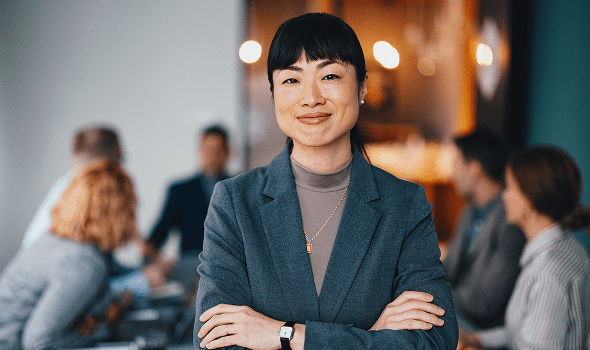 A person looks directly at the camera, crossing her arms and smiling while a group of people have a meeting in the background
