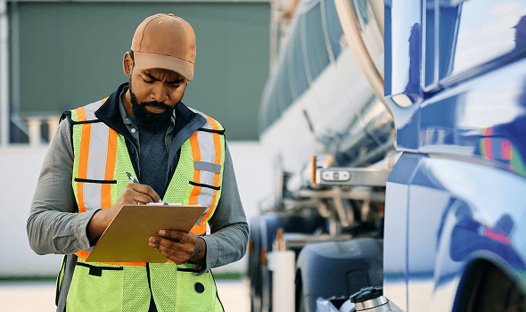 A worker writes on a clipboard while standing next to a tanker truck