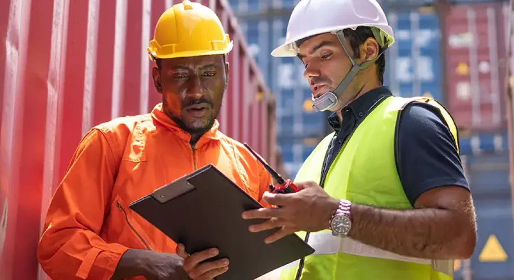 two men in safety gear reviewing a clipboard in a shipping yard